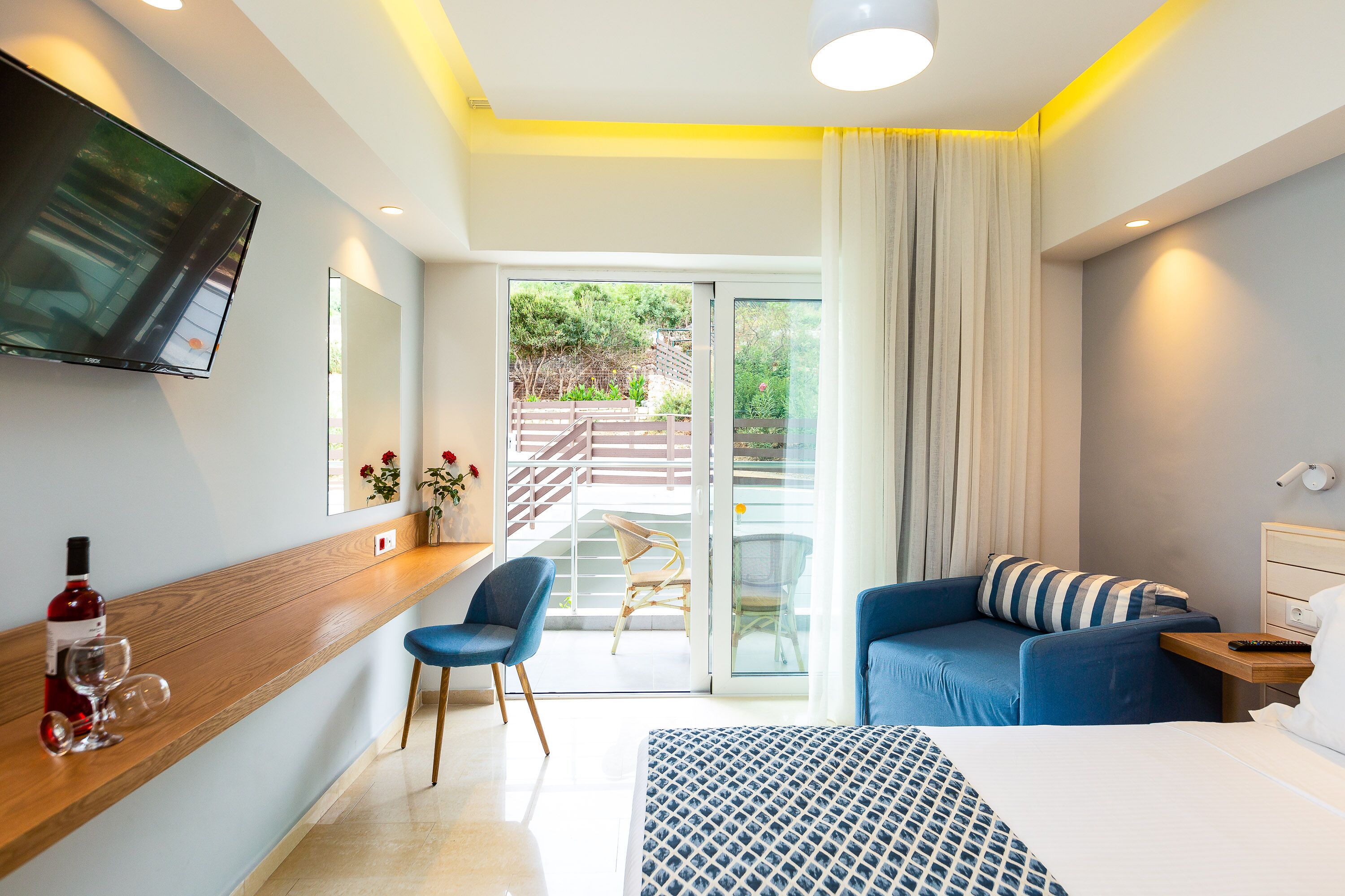 Una habitación de hotel moderna con una cama grande, un televisor montado en la pared, una mesita de madera con una botella de vino y copas, una silla azul y un balcón con vistas a la vegetación.