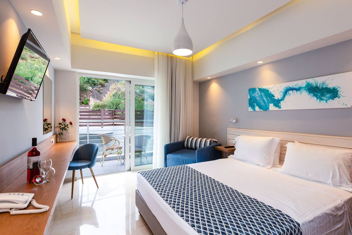 A modern hotel room with a large bed, a desk with a telephone and glasses, a chair, a TV mounted on the wall, and a balcony with a view of greenery.