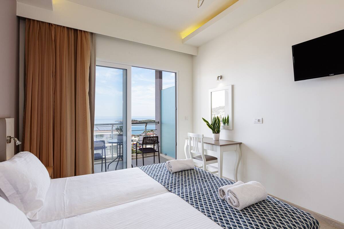 A hotel room with a bed, a desk, a chair, a TV, a balcony with a view, and a window with curtains.