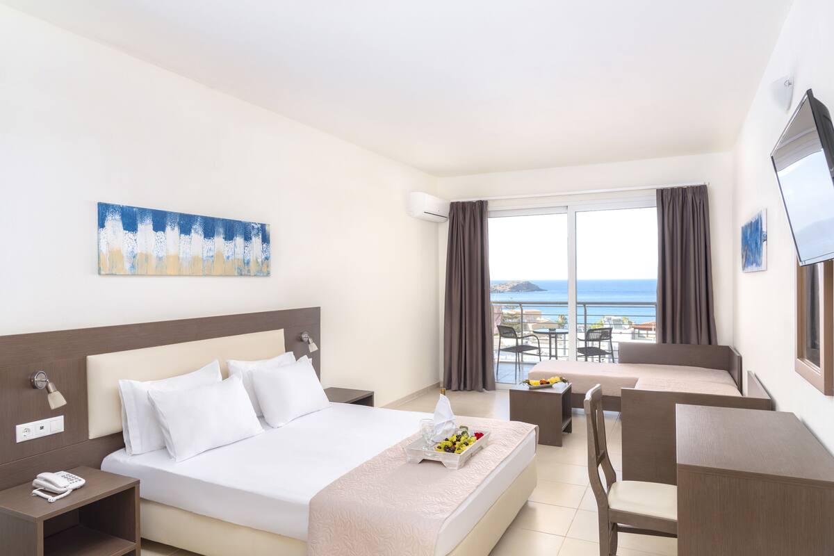 A hotel room with a bed, bedside table, desk, chair, and a balcony with a view of the sea.