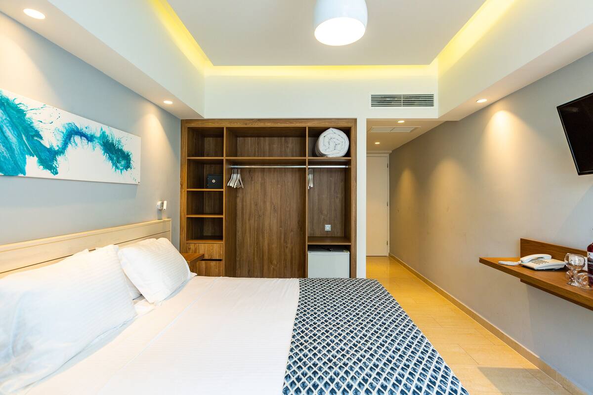 A hotel room with a bed, a wooden wardrobe, a desk with a phone and glasses, and a painting on the wall.