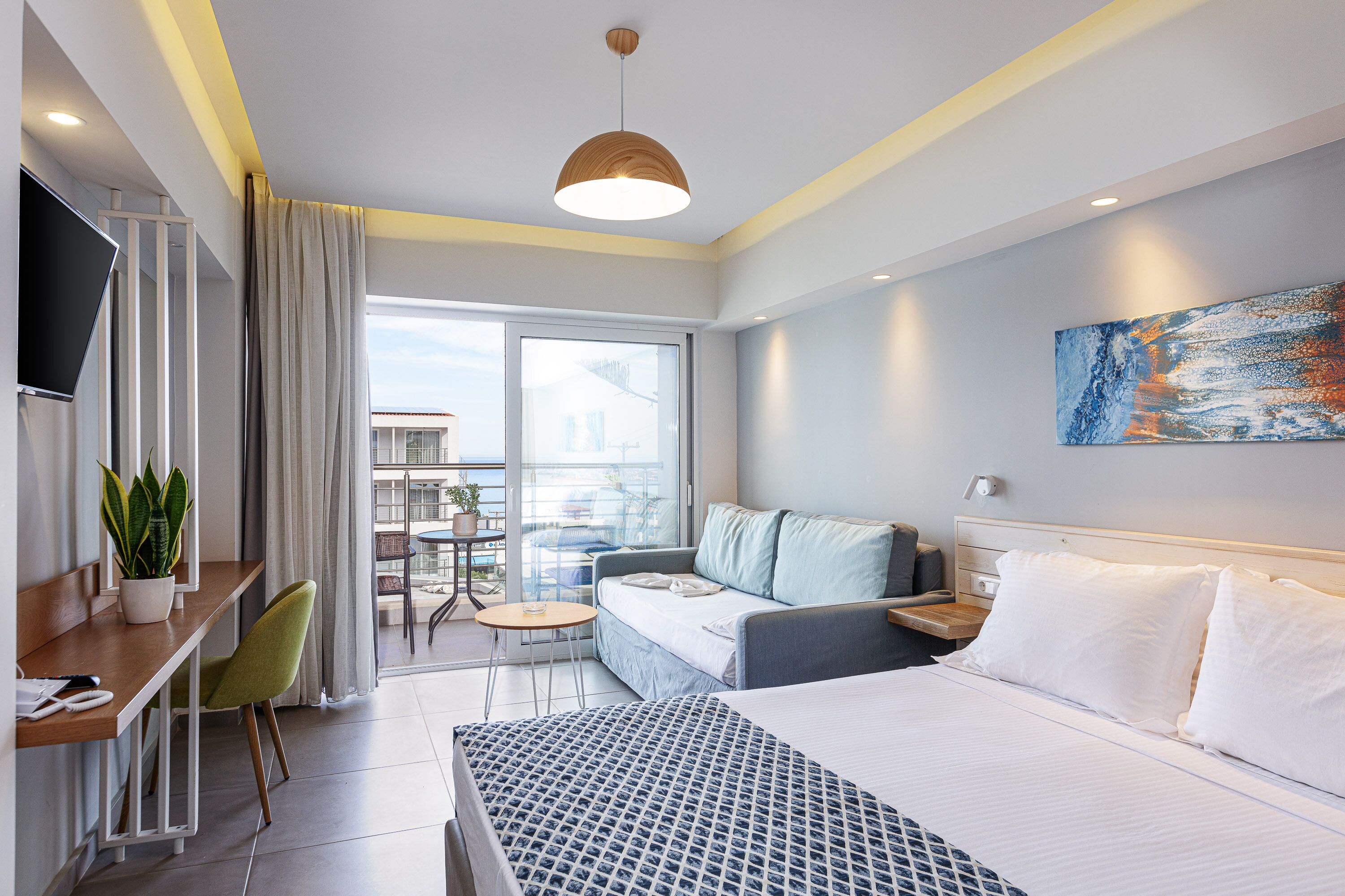 A modern hotel room with a bed, a desk, a chair, a sofa, and a balcony with a view.