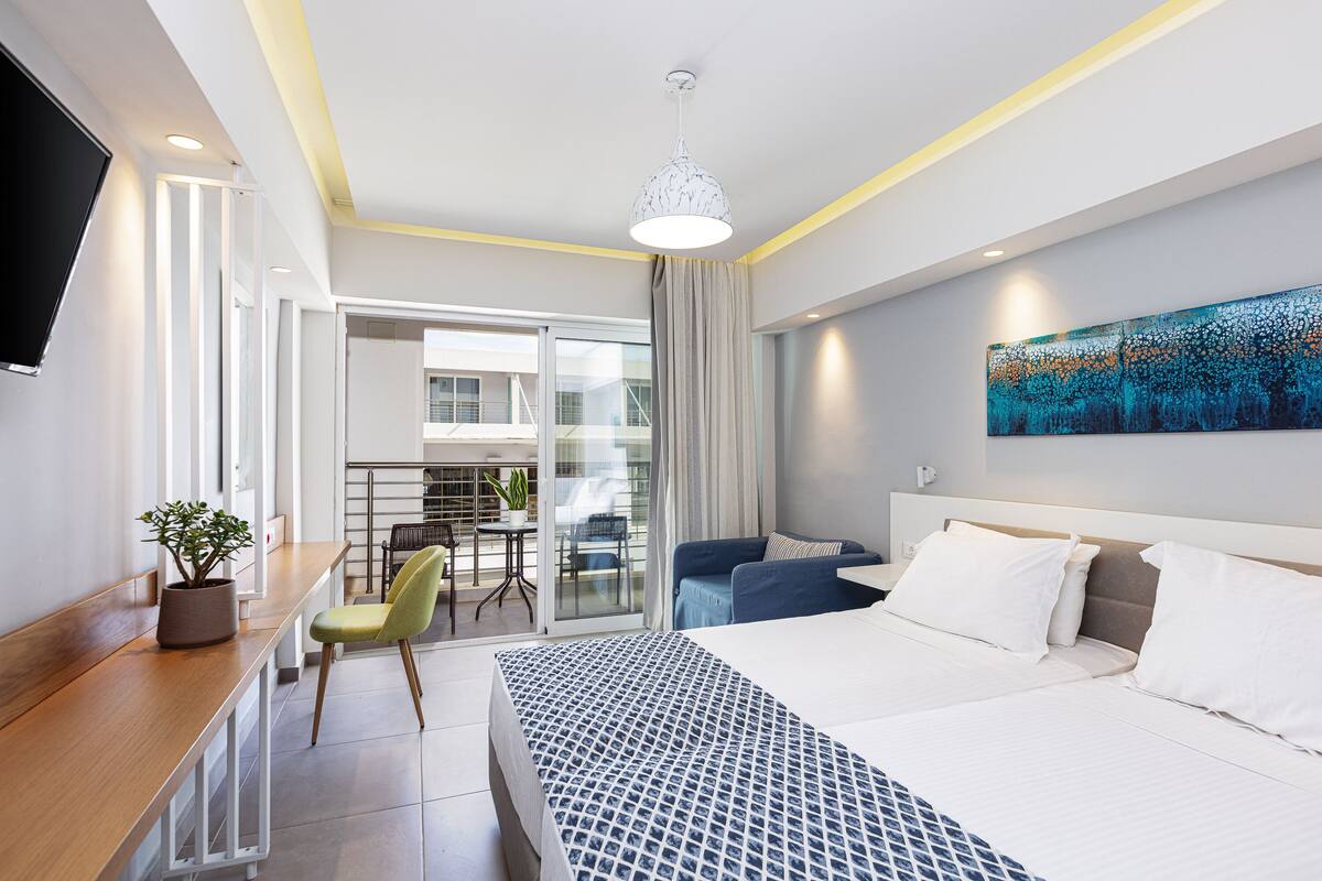 A modern hotel room with a large bed, a desk with a chair, a dining area with a table and chairs, and a balcony with a view.