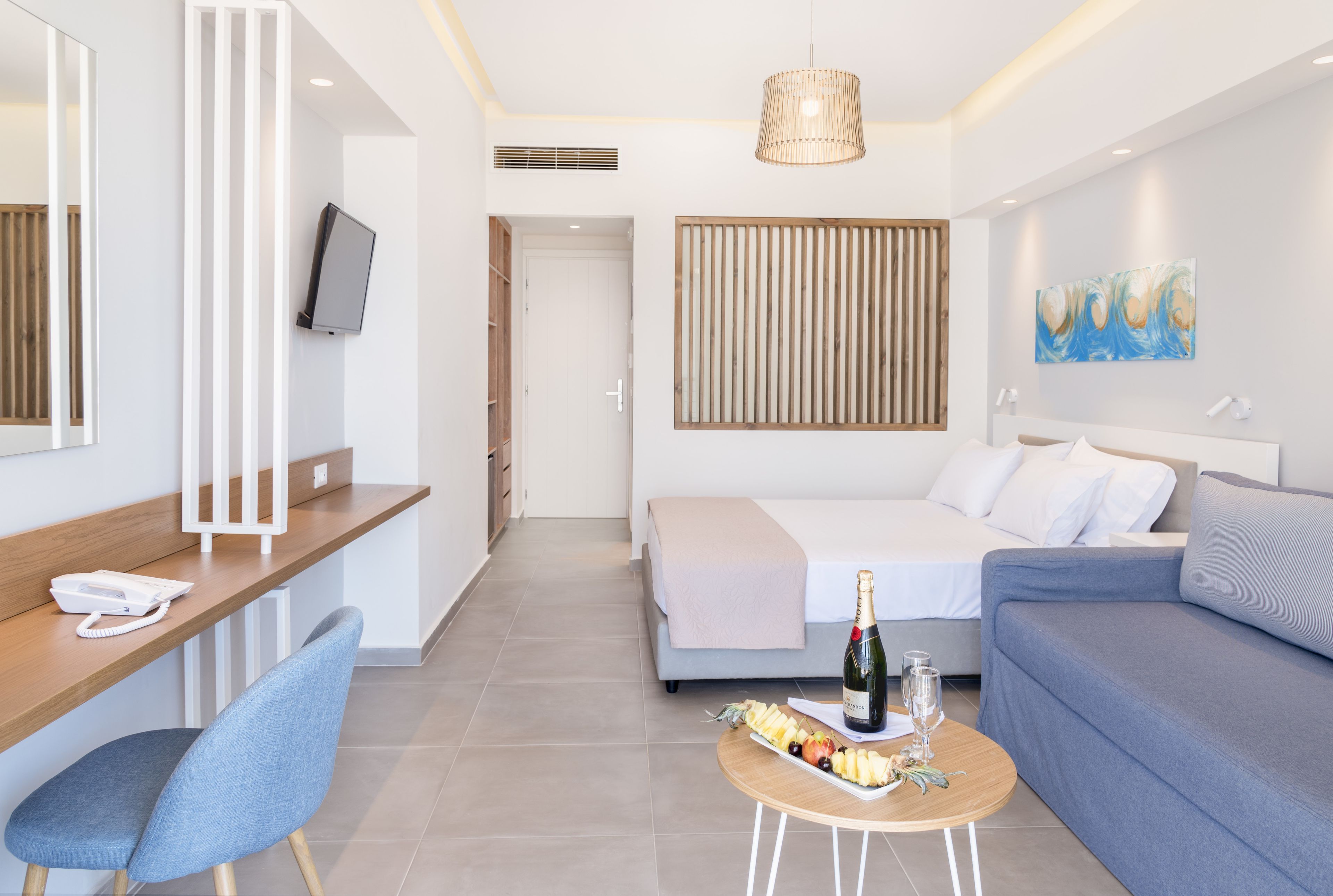 Una habitación de hotel moderna con cama, sofá, escritorio con teléfono, una pequeña mesa con comida y bebidas, televisor montado en la pared y un ventanal amplio.