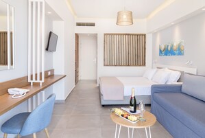 Una habitación de hotel moderna con cama, sofá, escritorio con teléfono, una pequeña mesa con comida y bebidas, televisor montado en la pared y un ventanal amplio.
