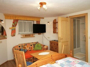 A cozy room with a wooden table, a sofa with colorful cushions, a television mounted on the wall, and a door leading to a bathroom.