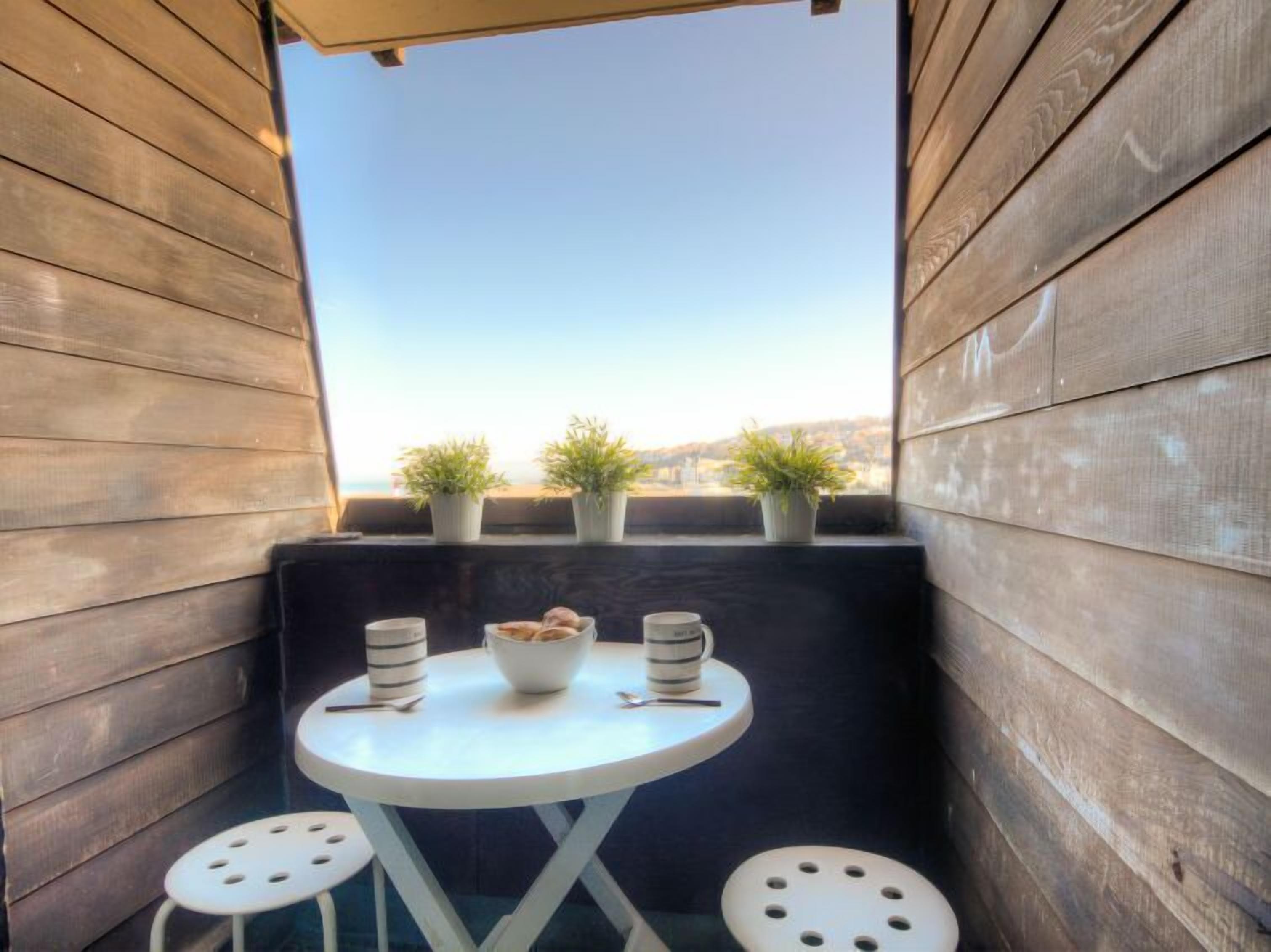 Un petit balcon avec des murs en bois, une table ronde, deux tabourets et trois plantes en pot.