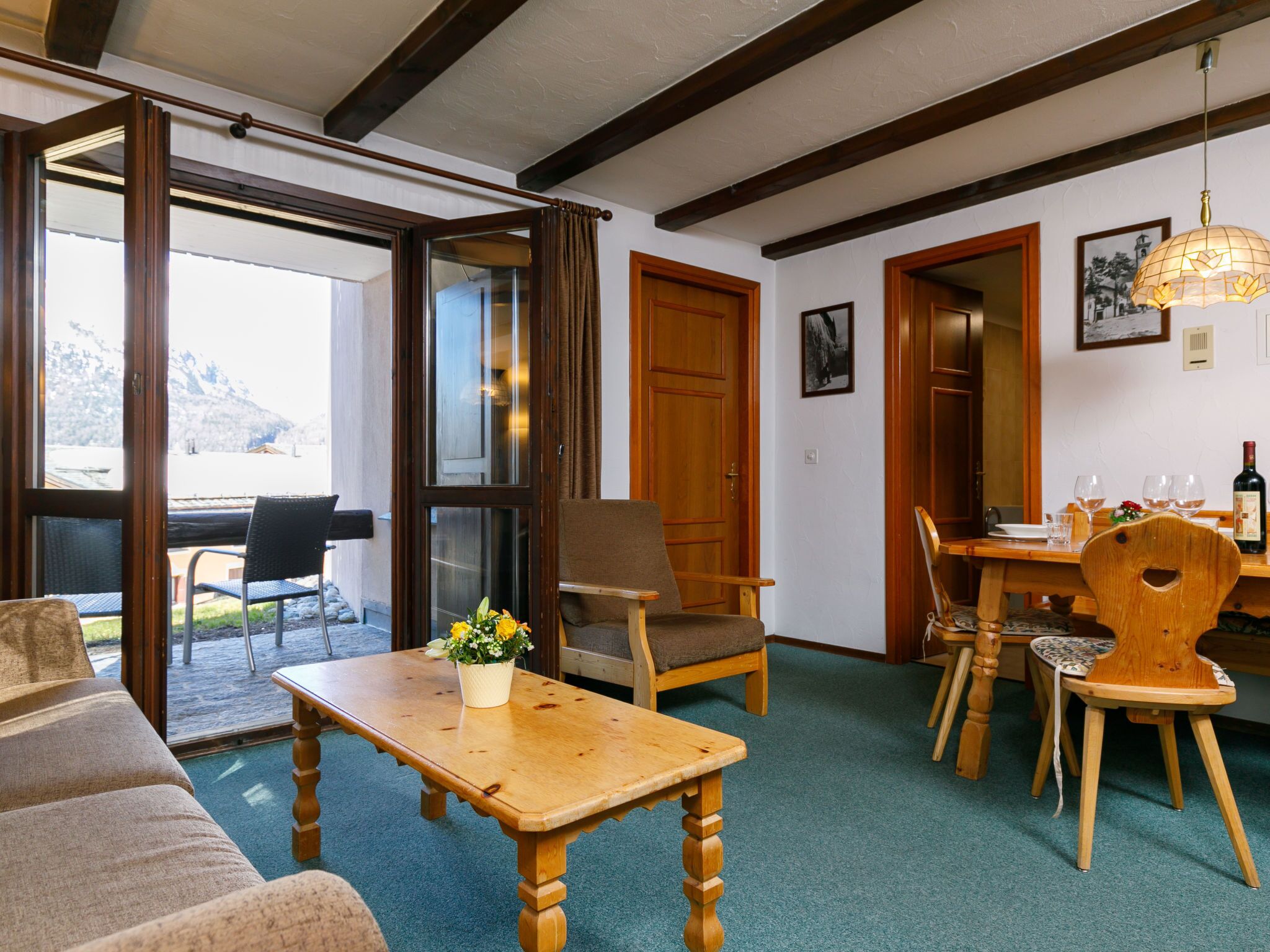 Una habitación acogedora con una mesa de madera, sillas y un sofá. Se ve una ventana con vistas a montañas.
