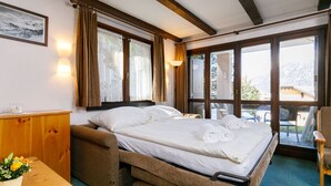 Un dormitorio con cama, un escritorio de madera, una silla, una mesita con una planta y una ventana con cortinas.