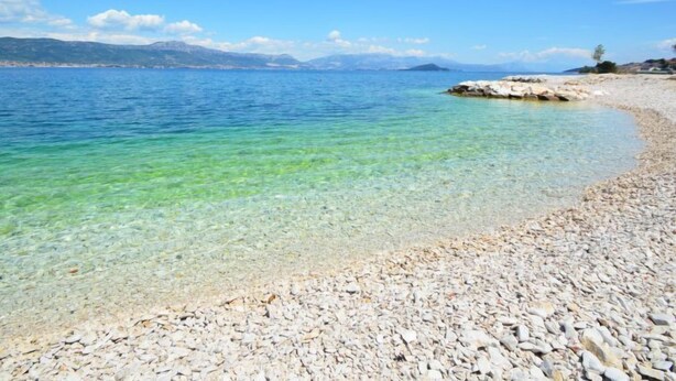 Une plage de galets avec une eau cristalline et des montagnes qui se dessinent à l’horizon.