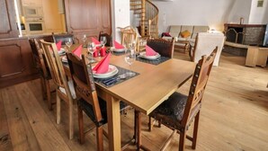 Un coin repas avec une table en bois dressée pour quatre personnes, entourée de chaises en bois. On aperçoit un escalier au fond, menant à un niveau supérieur.