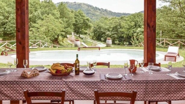 Un espacio de comedor al aire libre con mantel a cuadros, sillas de madera y vistas a una piscina y montañas.