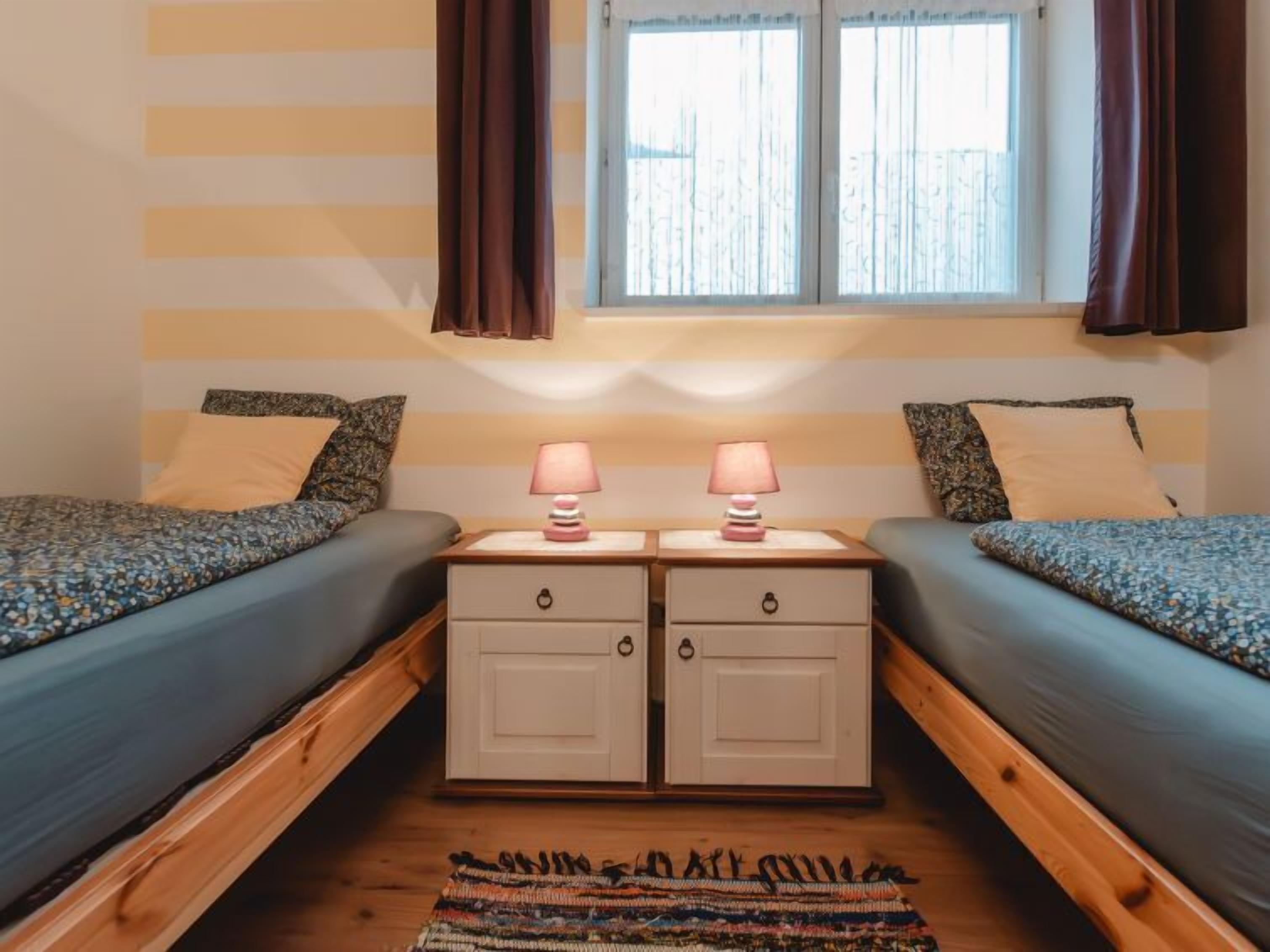 A room with two beds, each with a pillow and a lamp. There is a window with curtains, and a rug on the floor.