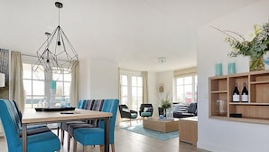 A modern living room with a dining area, featuring a geometric pendant light, a wooden coffee table, and a bookshelf with bottles. - Tapuit 8 by Interhome (Noordwijk)