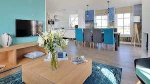 A modern living room with a flat-screen TV, a wooden coffee table with a vase of flowers, and a dining area with blue chairs and a chandelier. - Tapuit 8 by Interhome (Noordwijk)