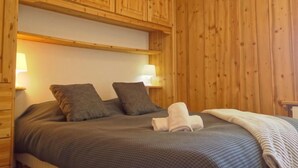 Quarto com painéis de madeira, uma cama, duas almofadas, uma toalha dobrada e duas luminárias fixadas na parede.