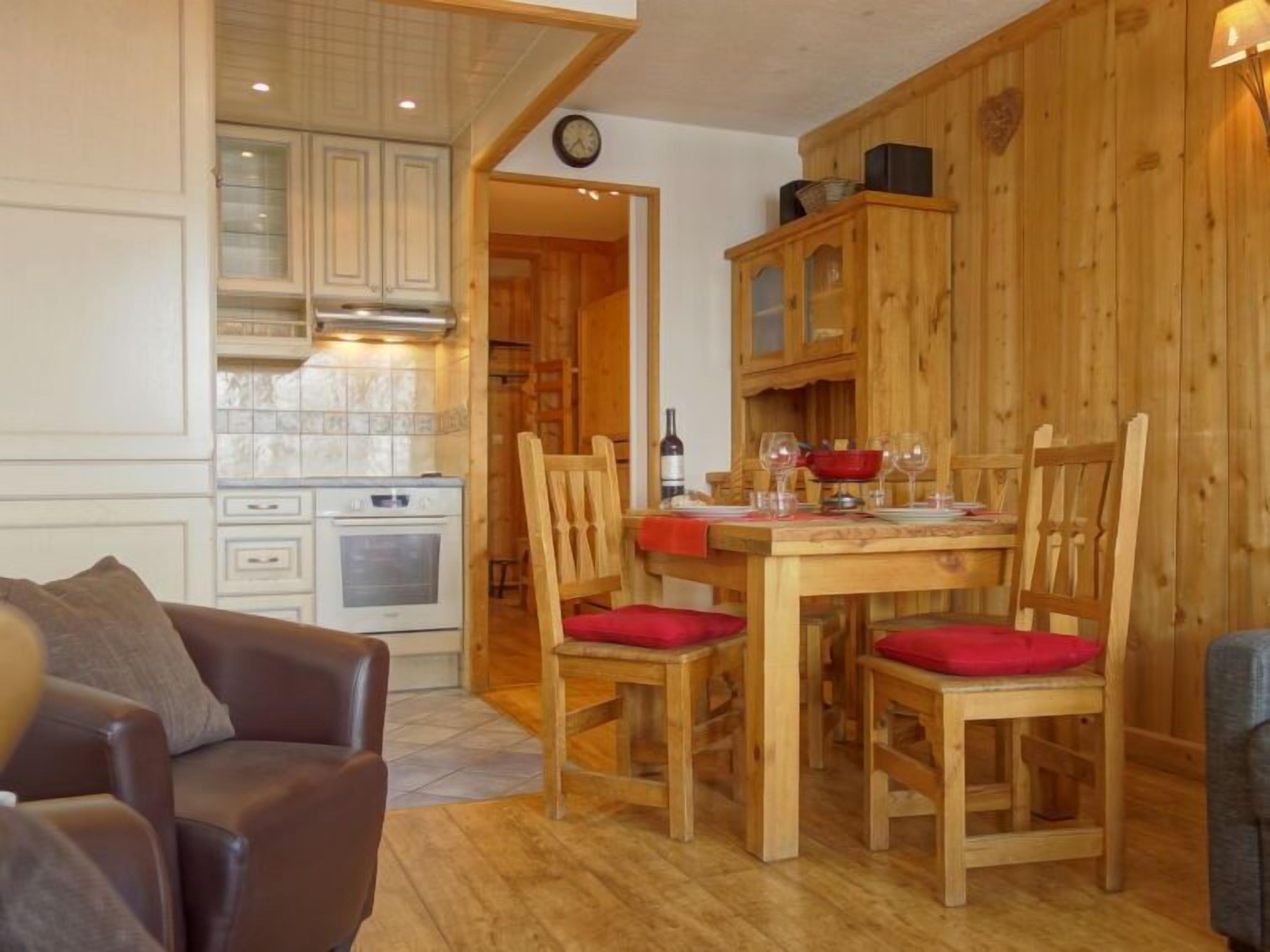 Un salon chaleureux avec un canapé en cuir marron et une table à manger en bois, mise pour quatre personnes, agrémentée de coussins rouges. On aperçoit au fond une cuisine avec des meubles blancs et une cuisinière.