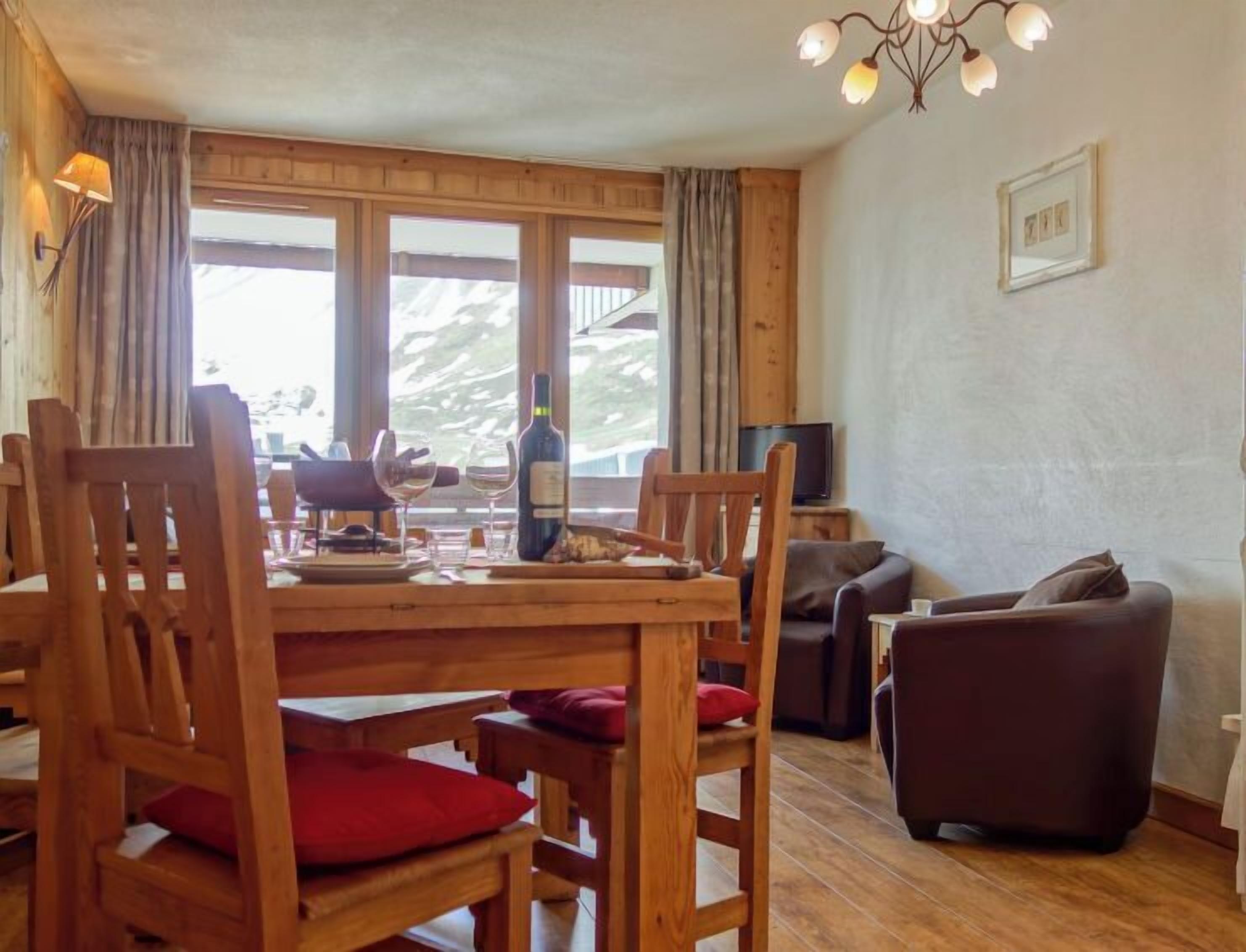 Ein Holztisch mit Stühlen, eine Weinflasche und Gläser. Ein Fernseher steht auf einem kleinen Tisch neben dem Essbereich.