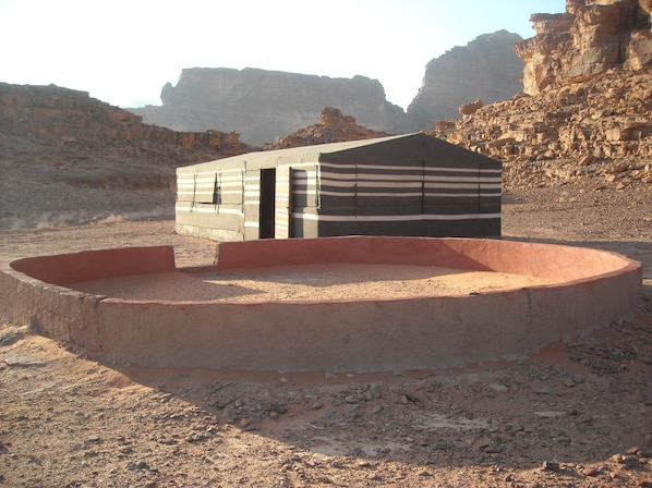 A caravan-style building with a flat roof and striped awning, situated in a desert with rocky terrain and cliffs in the background.