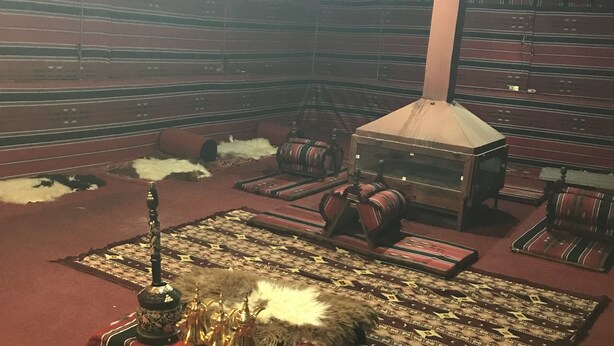A room with a high ceiling, patterned walls, and a central fireplace. There are several sofas and rugs arranged around the fireplace. - Bedouin expedition Camp (Wadi Rum)