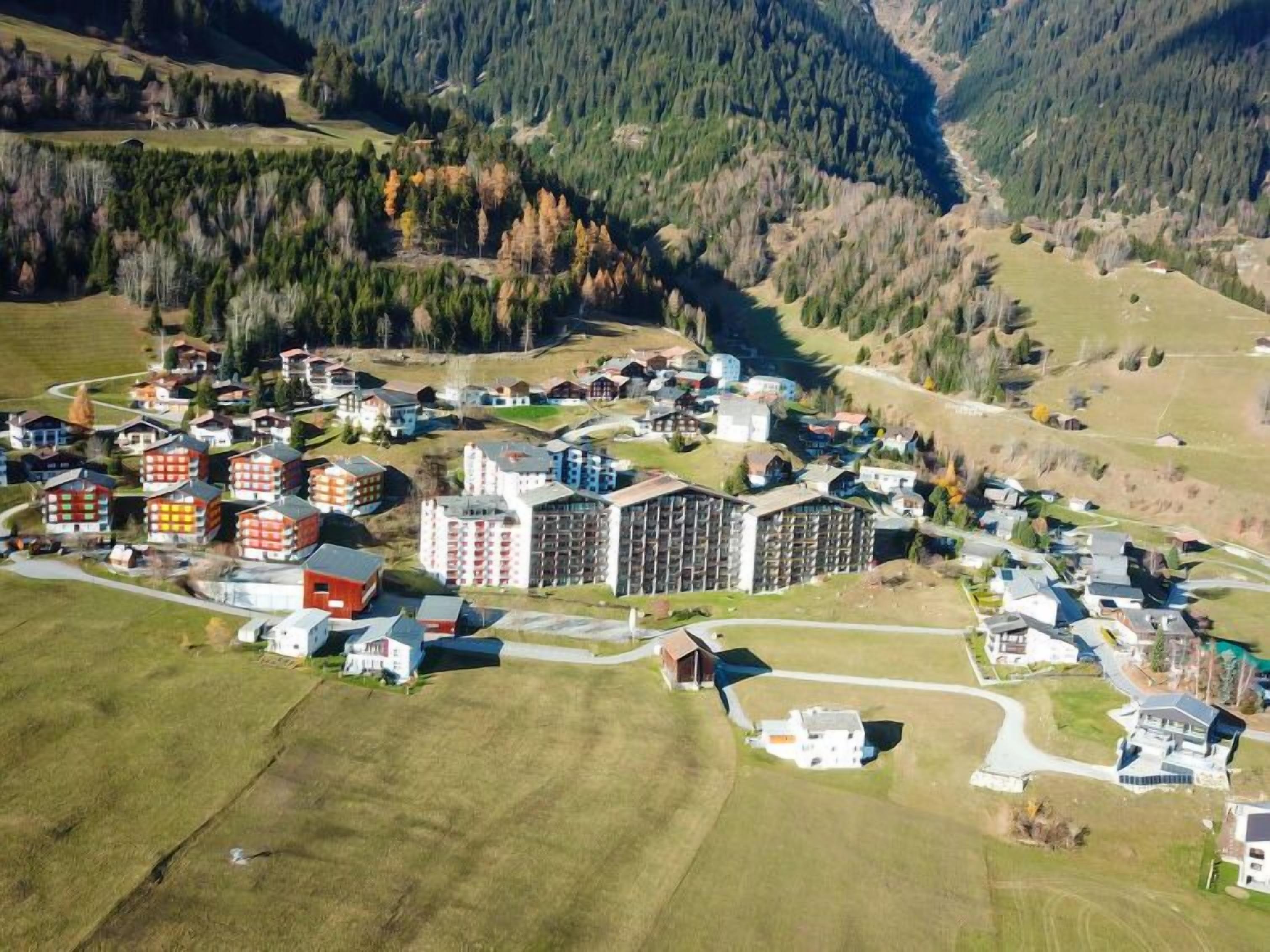An aerial view of a residential area with a mix of single and multi-story buildings, surrounded by forested hills and open fields.