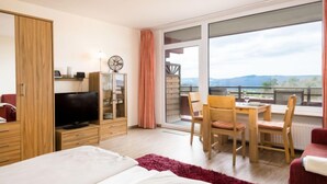 A modern hotel room with a large bed, a TV, a wooden cabinet, a balcony with a table and chairs, and a view of the mountains. - Eifelblick (B906) by Interhome (Lahnstein (Koblenz))