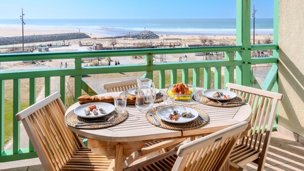 Een houten tafel met borden eten, een karaf en glazen op een balkon met uitzicht op een strand.