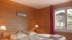 A wooden room with a bed, two bedside lamps, a window with a red curtain, and a picture on the wall. - Sulegg Apt. I by Interhome (Wengen)