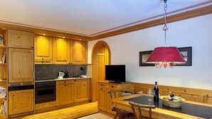 A kitchen with wooden cabinets, a dining table, chairs, a TV, and a red pendant light. - Residenza Chesa Margun 57-4 by Interhome (Silvaplana-Surlej)