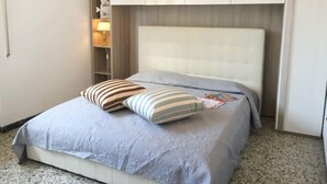 A neatly made bed with a light blue bedspread and two striped pillows, a bedside lamp, and a white wardrobe above the bed. - Vacation home Sandra by Interhome (Forte dei Marmi)