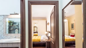 Habitación de hotel con piso de madera, cama con ropa de cama amarilla, mesita de noche con lámparas, espejo y baño con lavamanos y ducha.