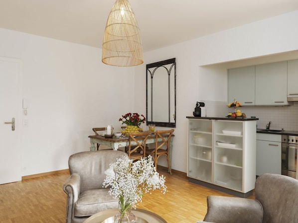 A modern living room with a sofa, a dining table with chairs, a kitchen area with cabinets and a stove, and a hanging light fixture.