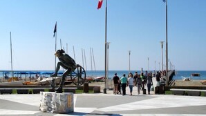 Una plaza pública con una estatua, banderas y un paseo marítimo junto al mar.