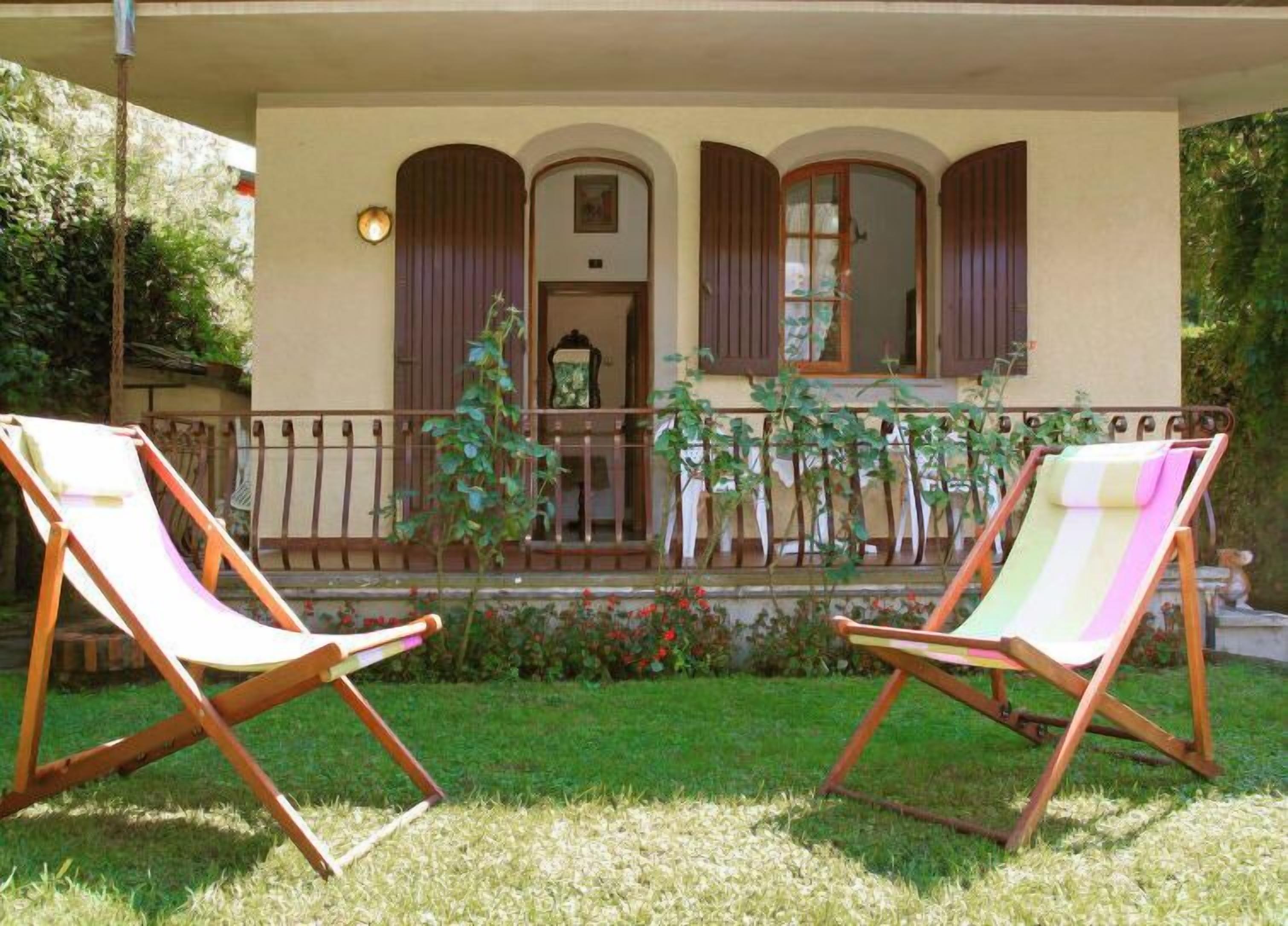 A two-story building with a balcony, two wooden lounge chairs, and a garden with flowers and plants.