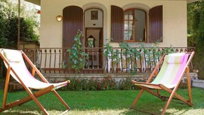 Un edificio de dos pisos con un balcón, dos sillones de madera y un jardín con flores y plantas.