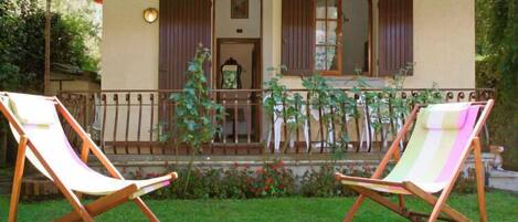 A two-story building with a balcony, two wooden lounge chairs, and a garden with flowers and plants.