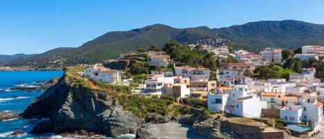 A coastal village with white buildings, rocky coastline, and mountains in the background.
