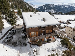 Una cabaña de madera con balcón, rodeada de montañas nevadas y un área de estacionamiento.