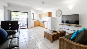 A modern living room with a sofa, coffee table, and TV. It includes a kitchen with wooden cabinets and a dining area with chairs. - THE REGAL - APARTMENT WITH SPA BATH - PRIME LOCATION IN TOWN CENTRE (Port Douglas)