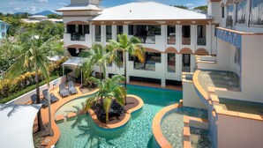 A resort with a central pool surrounded by palm trees and lounge chairs, a multi-story building with balconies, and a mountainous backdrop. - THE REGAL - APARTMENT WITH SPA BATH - PRIME LOCATION IN TOWN CENTRE (Port Douglas)