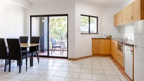 A modern kitchen with wooden cabinets, a built-in oven, and a dishwasher. There is a dining area with a table and chairs, and a sliding glass door leading to a balcony. - THE REGAL - APARTMENT WITH SPA BATH - PRIME LOCATION IN TOWN CENTRE (Port Douglas)