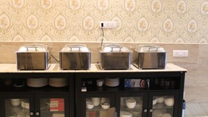 A buffet setup with stainless steel chafing dishes on a marble countertop, surrounded by glass cabinets containing plates and bowls. - Royal Residence Huda City Centre (Gurugram)