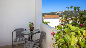 A balcony with wicker chairs and a small table, overlooking a hillside with buildings. - Apartments Milka (Hvar)
