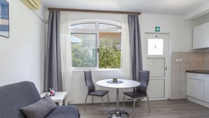 A compact living space with a sofa, a small table with a cup, two chairs, a window with curtains, a door, and a kitchenette. - Apartments Milka (Hvar)