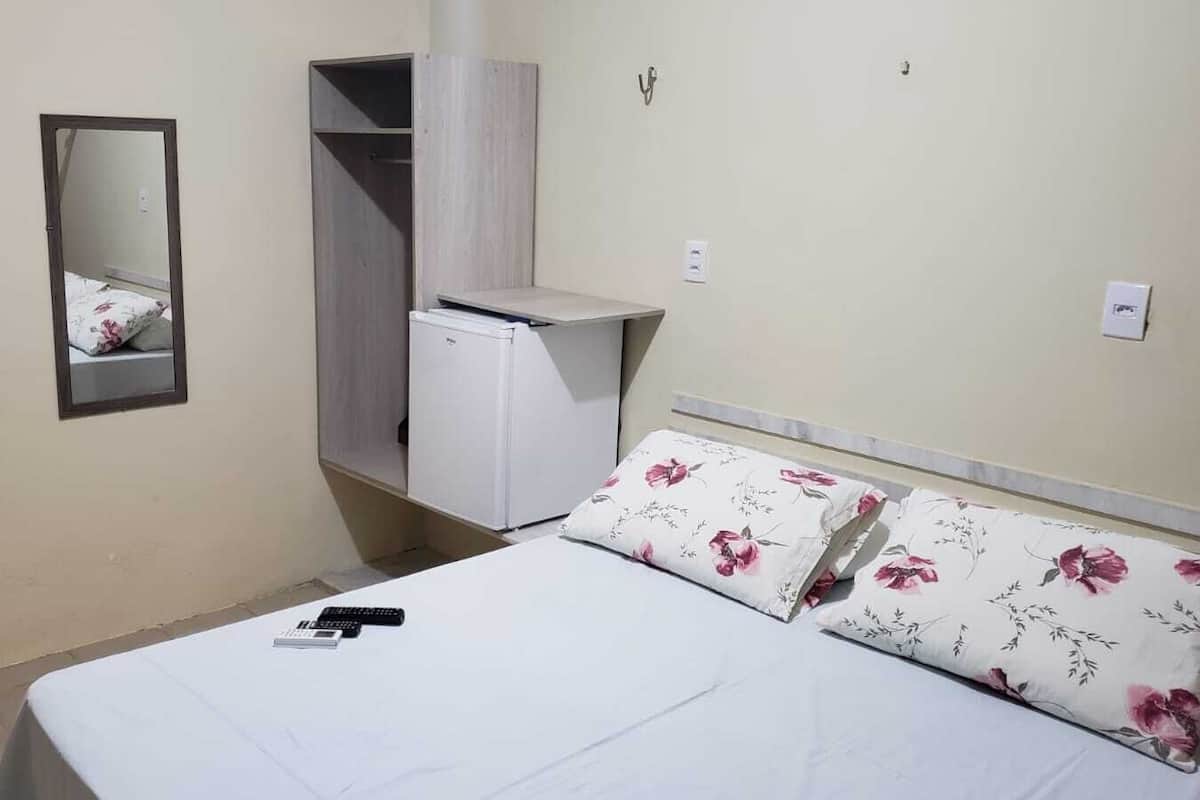 Quarto pequeno e bem organizado, com cama, guarda-roupa, espelho e um pequeno eletrodoméstico branco, provavelmente uma geladeira.
