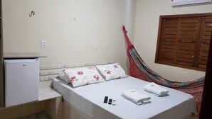 Un lit simple avec une literie à motifs floraux, un petit réfrigérateur blanc, un hamac rouge et une fenêtre avec des volets en bois.