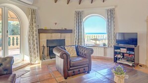 A living room with a fireplace, a sofa, a television, and a view of the sea. - Villa Bellevue by Interhome (Jávea)
