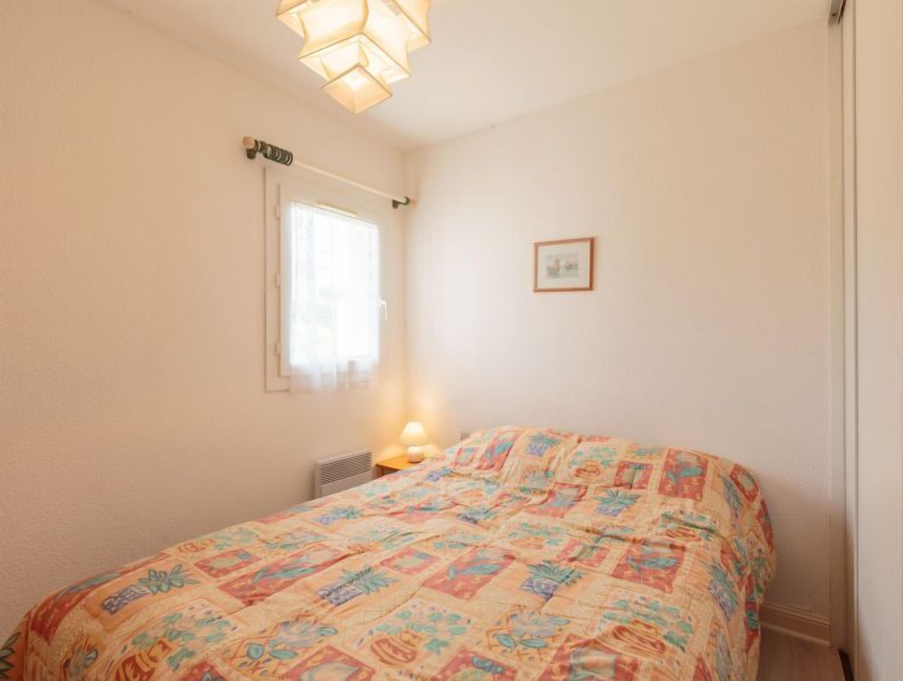 Une chambre à coucher avec un lit, une fenêtre, un tableau accroché au mur et un luminaire fixé au plafond.