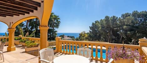A terrace with a round table and chairs, overlooking a pool and the sea.