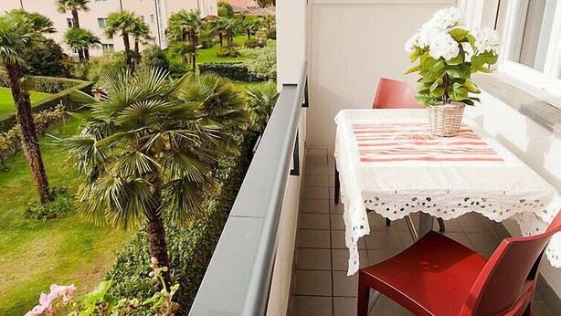 A balcony with a table and chairs, overlooking a garden with palm trees and a view of houses in the distance. - Double Room by Interhome (Ascona)